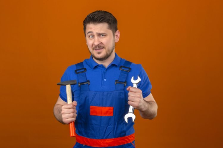 builder man wearing construction uniform and security helmet holding wrench and hammer in hands looking at camera with smile on face standing over isolated orange background